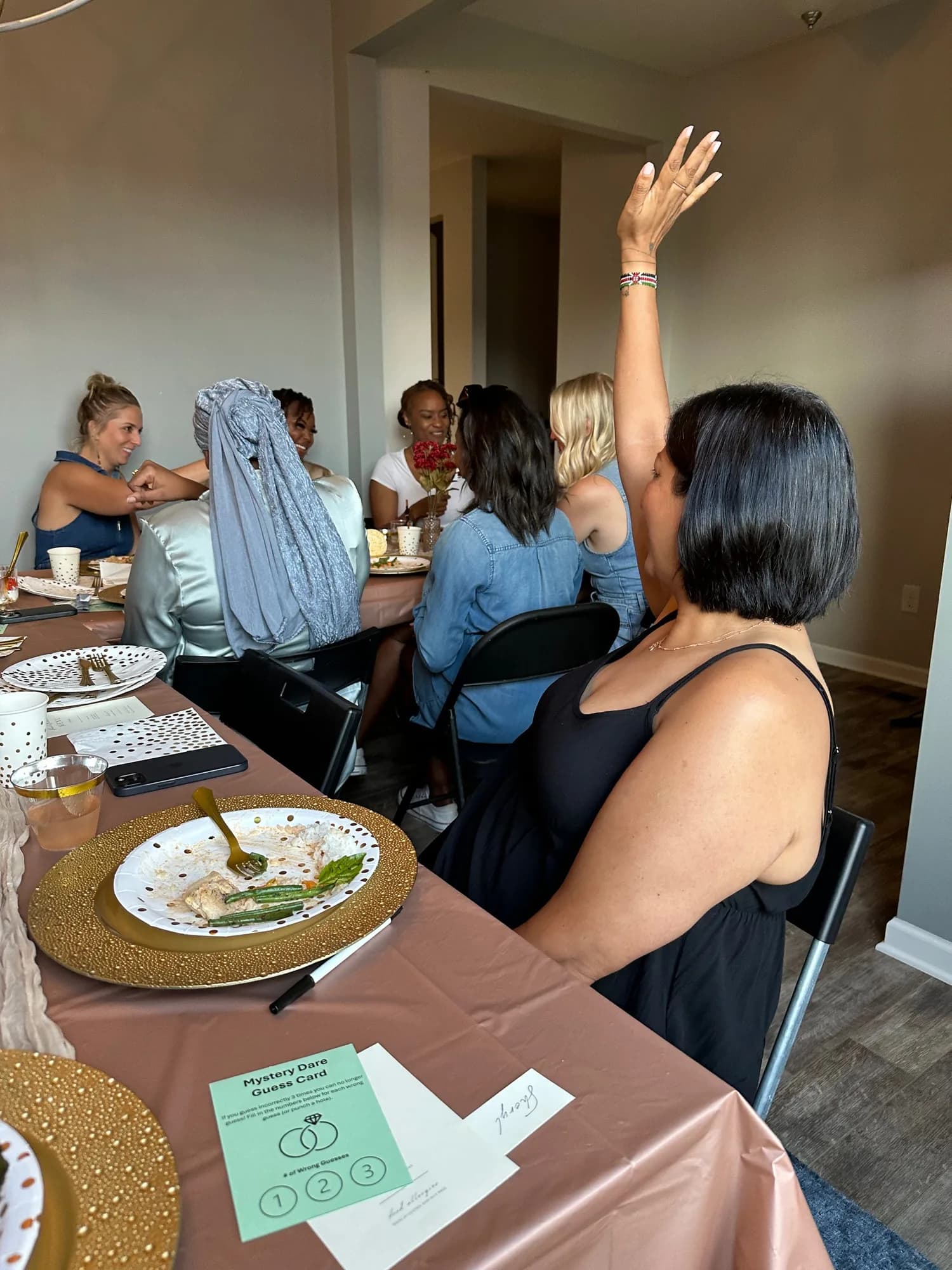 A woman raising her hand to make a guess during a dinner party, a Mystery Dare guess card on the table